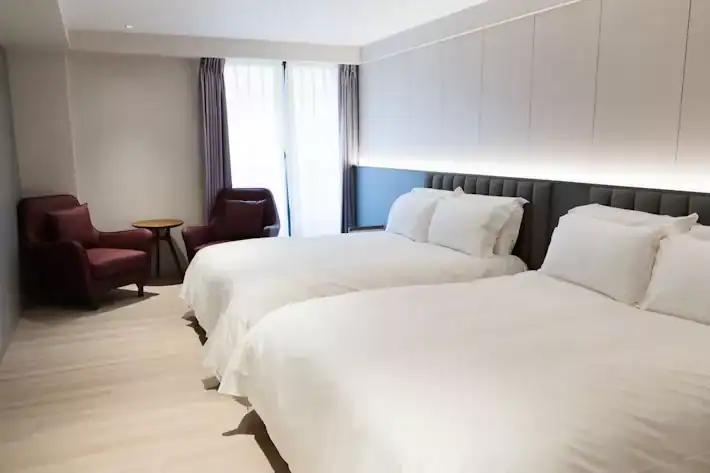 塔木德酒店雙床客房，配置兩張大床與簡約木質地板，窗邊設有兩張深紅色單椅與小圓桌，柔和燈光與灰白色牆面營造出明亮舒適的住宿氛圍。
