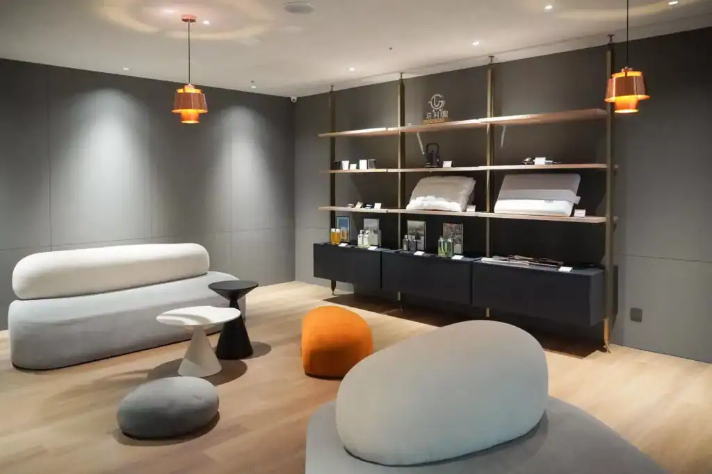 塔木德酒店展示空間，採用極簡灰色調與木質地板設計，擺放圓潤造型的沙發與圓形坐墊，橘色吊燈散發柔和光線，牆面層架陳列枕頭、寢具與香氛用品，展現舒適又有質感的氛圍。