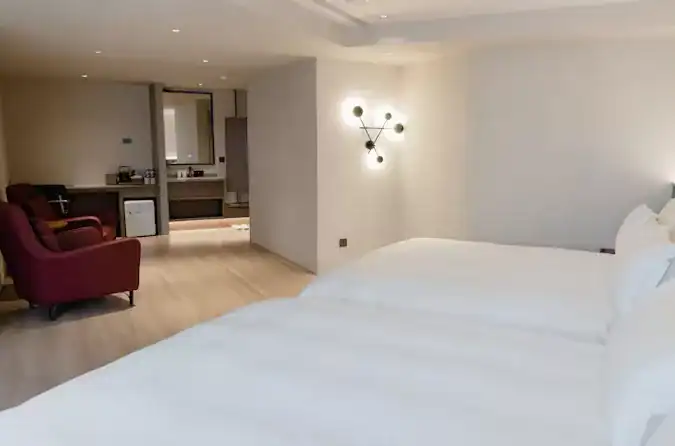 塔木德酒店寬敞雙床客房，採用柔和燈光與極簡設計風格，房內設有雙人床、紅色單椅與迷你吧台，整體空間明亮舒適，展現現代感與放鬆氛圍。