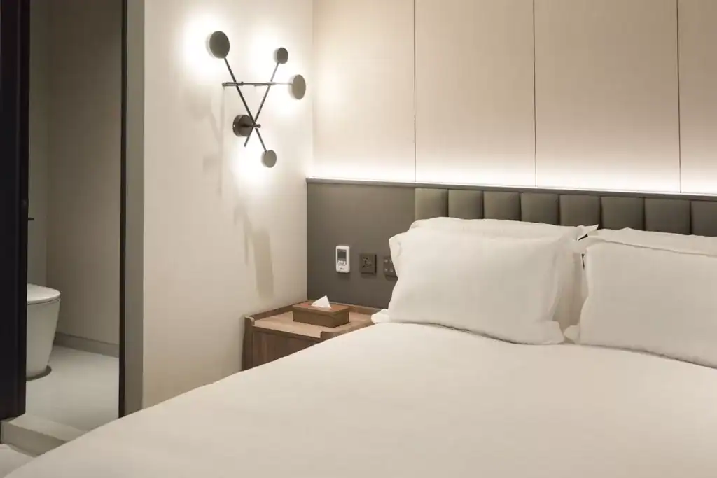 塔木德酒店台中台灣大道館客房一隅，潔白床鋪與柔和燈光營造溫馨氛圍，床頭設有深灰軟墊與木質床邊櫃，牆上裝有幾何造型壁燈，左側可見衛浴入口。