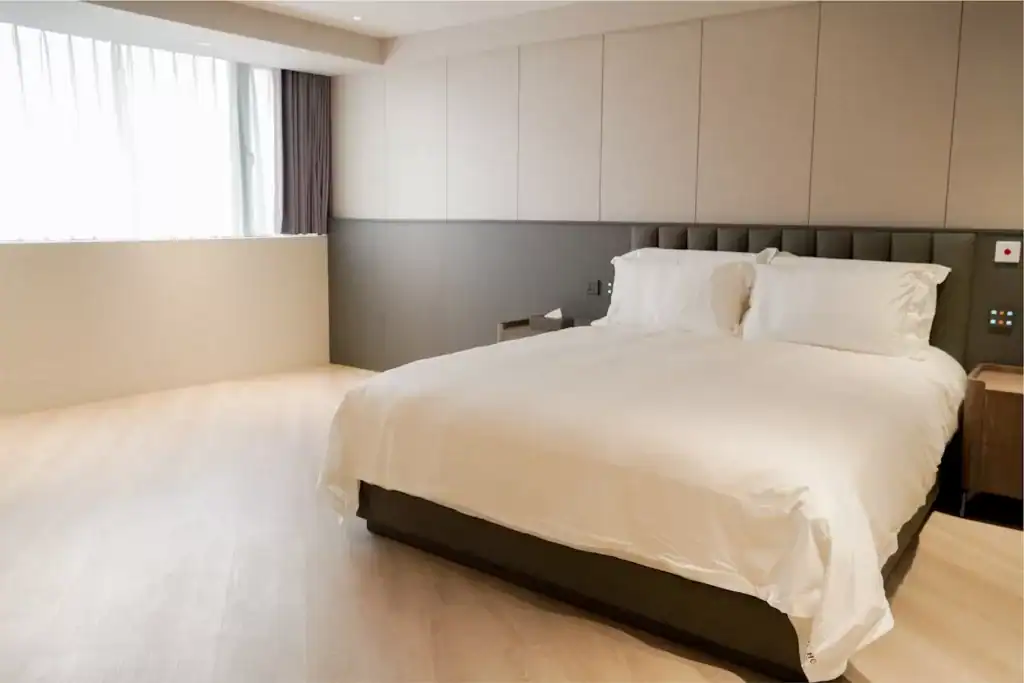 塔木德酒店台中台灣大道館無障礙客房，設有一張大床與柔軟白色寢具，房間以淺木地板與灰米色牆面呈現簡約風格，大片窗戶引入自然光，空間寬敞明亮、動線平整無障礙。