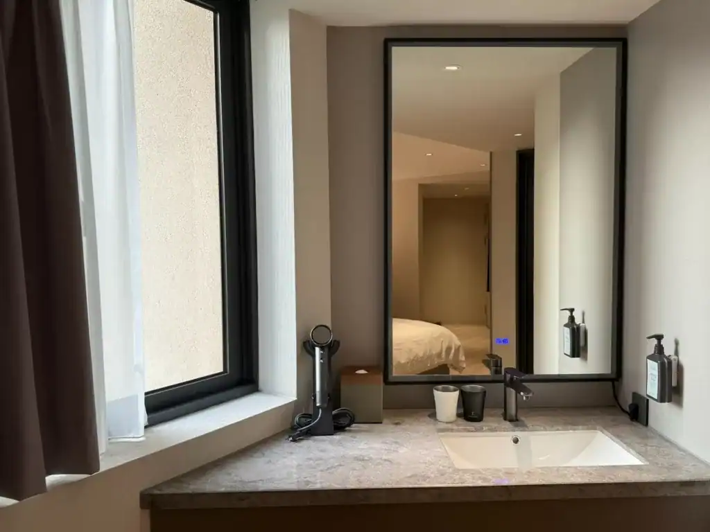 塔木德酒店台中台灣大道館客房內的化妝台空間，設有大面鏡子與洗手台，桌面上擺放吹風機、紙巾盒與兩個馬克杯，整體採簡約現代風格，窗邊自然光灑入，營造明亮舒適的住宿氛圍。