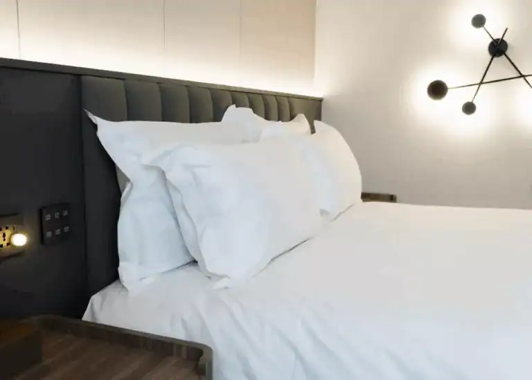 現代風格的飯店房間一隅，特寫展示潔白柔軟的雙人床與多顆蓬鬆枕頭，床頭板設有插座與閱讀燈，背景為簡約牆面與幾何造型壁燈，營造出舒適放鬆的住宿氛圍。